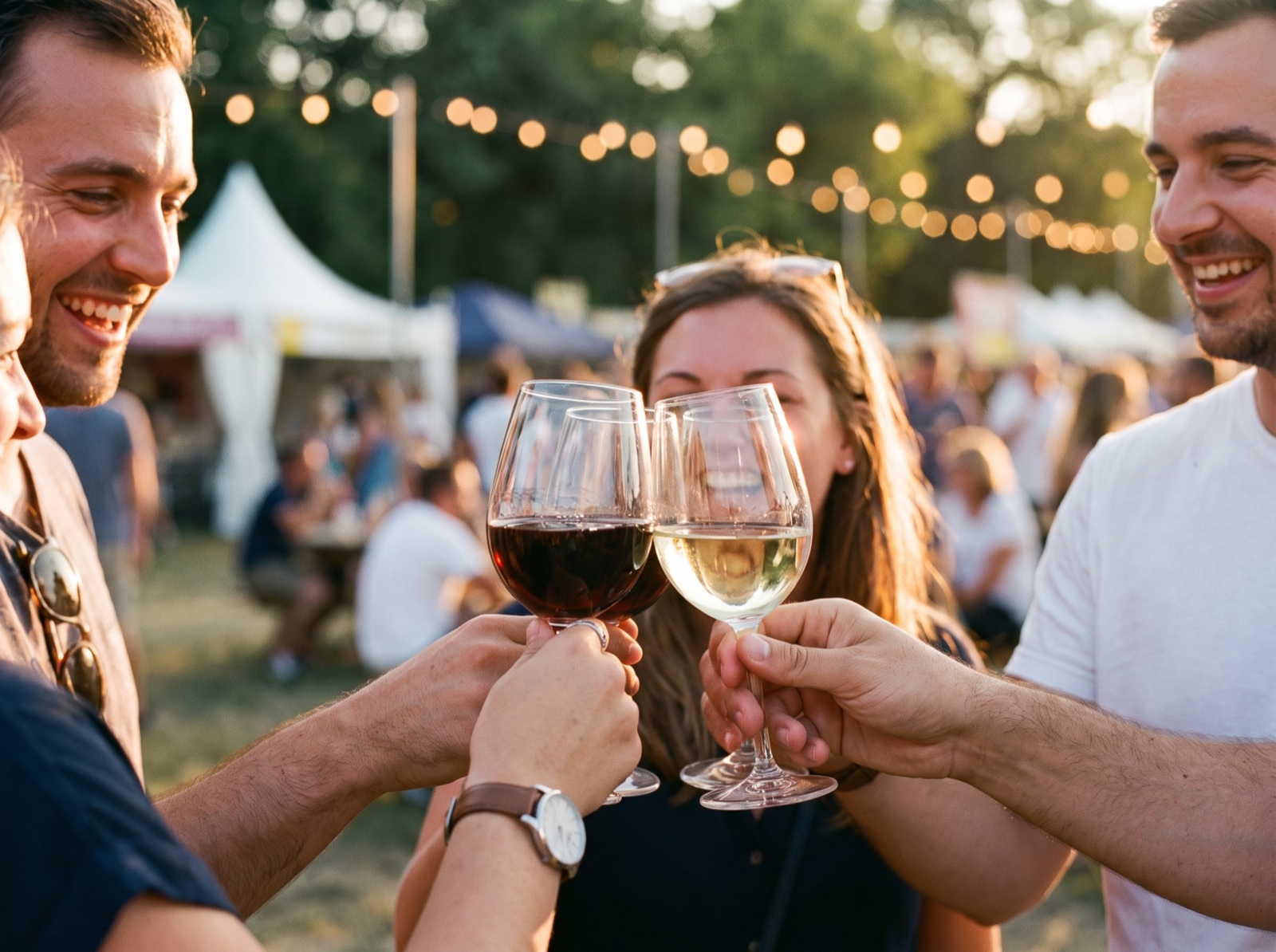 Vier Freunde stoßen mit Rot- und Weißwein an, im Hintergrund Zelte und Lichterketten